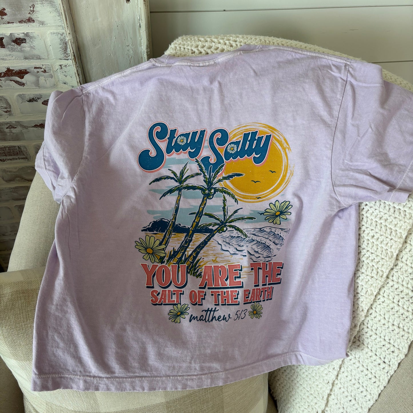 Lilac-colored t-shirt with 'Stay Salty' text and a palm tree design on the front, placed on a chair with a white knit blanket.