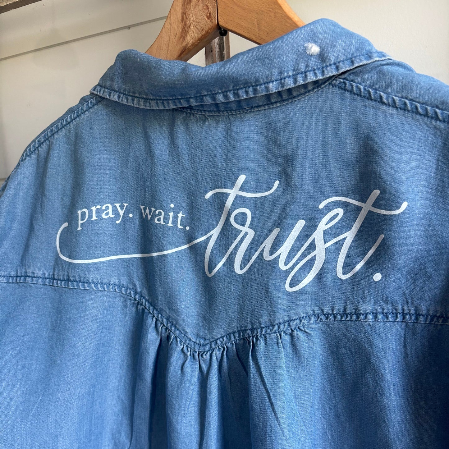 Blue denim shirt with white text 'pray. wait. trust.' on back