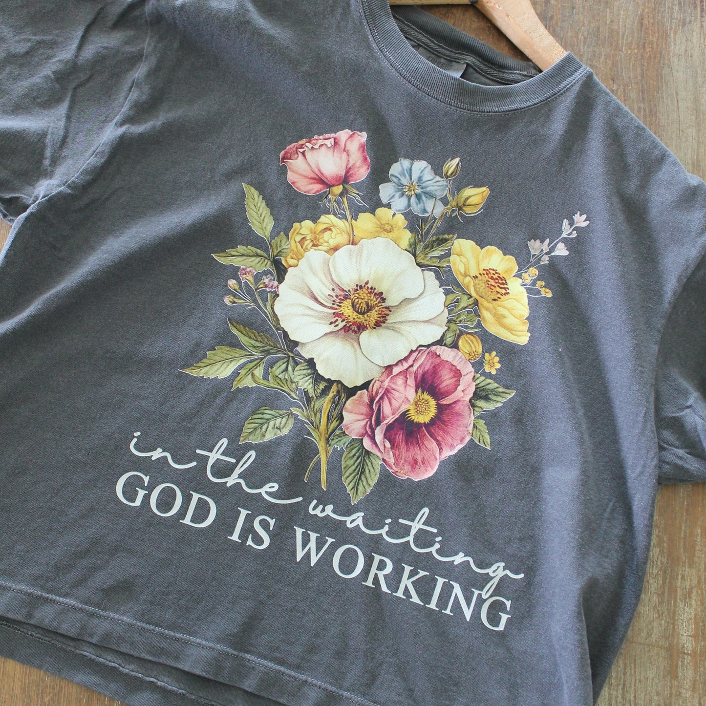 A gray cropped t-shirt with a floral design and the text 'In the waiting, God is working' printed on the front.